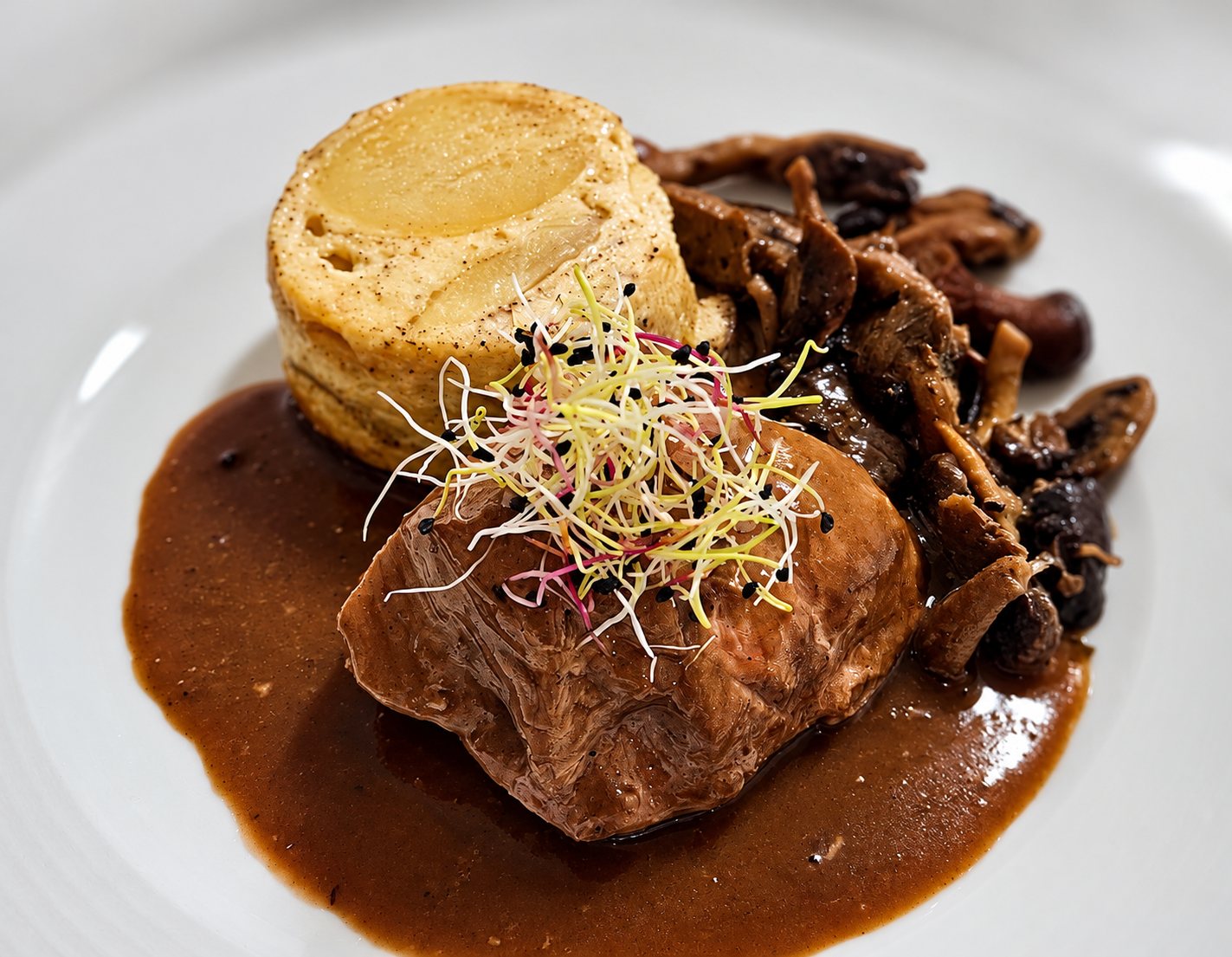 Une assiette blanche présente un morceau de viande nappé de sauce brune, accompagné de champignons et d'un petit flan, le tout décoré de jeunes pousses colorées.