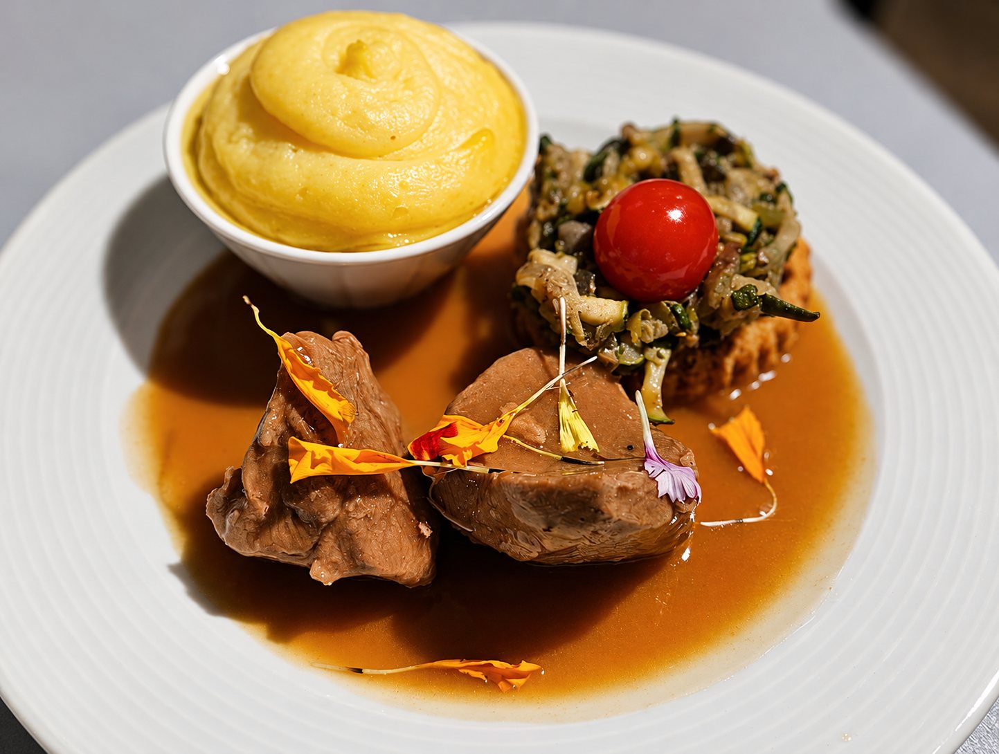 Assiette blanche garnie de deux morceaux de viande nappés de sauce brune, d'une purée jaune en ramequin et de légumes surmontés d'une tomate cerise, décorée de pétales de fleurs comestibles.