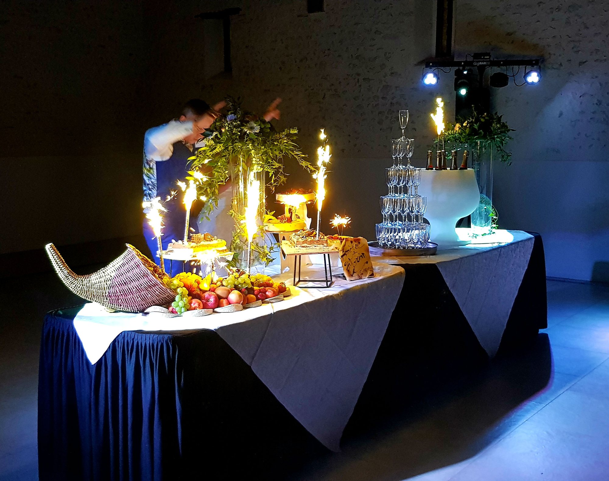 Une longue table de réception drapée de bleu nuit et de blanc, avec une corne d'abondance de fruits, une pyramide de coupes, des gâteaux et des bougies étincelantes, dans une salle sombre.