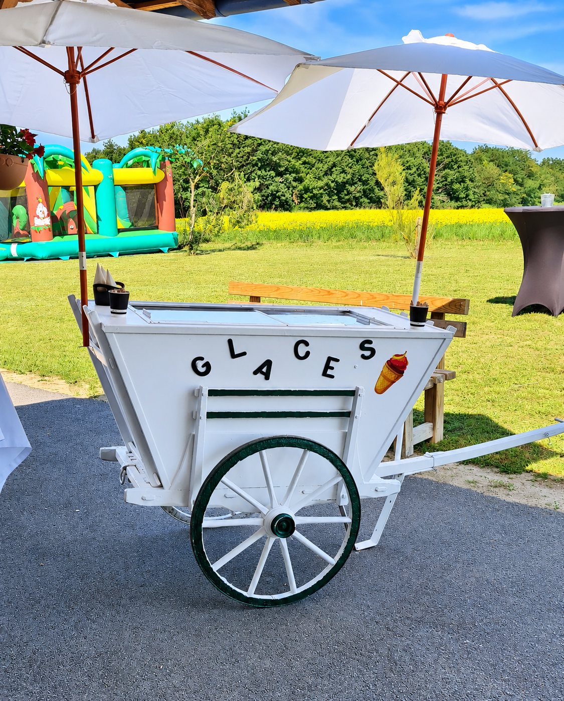 Une charrette à glaces blanche en bois, ornée du mot GLACES, est installée sous deux parasols devant une pelouse et un château gonflable.