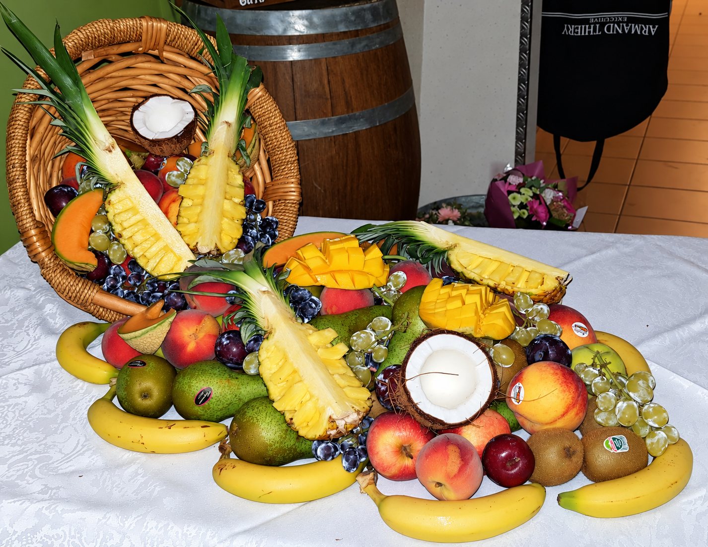 Une corbeille en osier renversée laisse s'échapper des ananas, une mangue, des noix de coco, des bananes, des poires, des pêches et du raisin sur une nappe blanche.