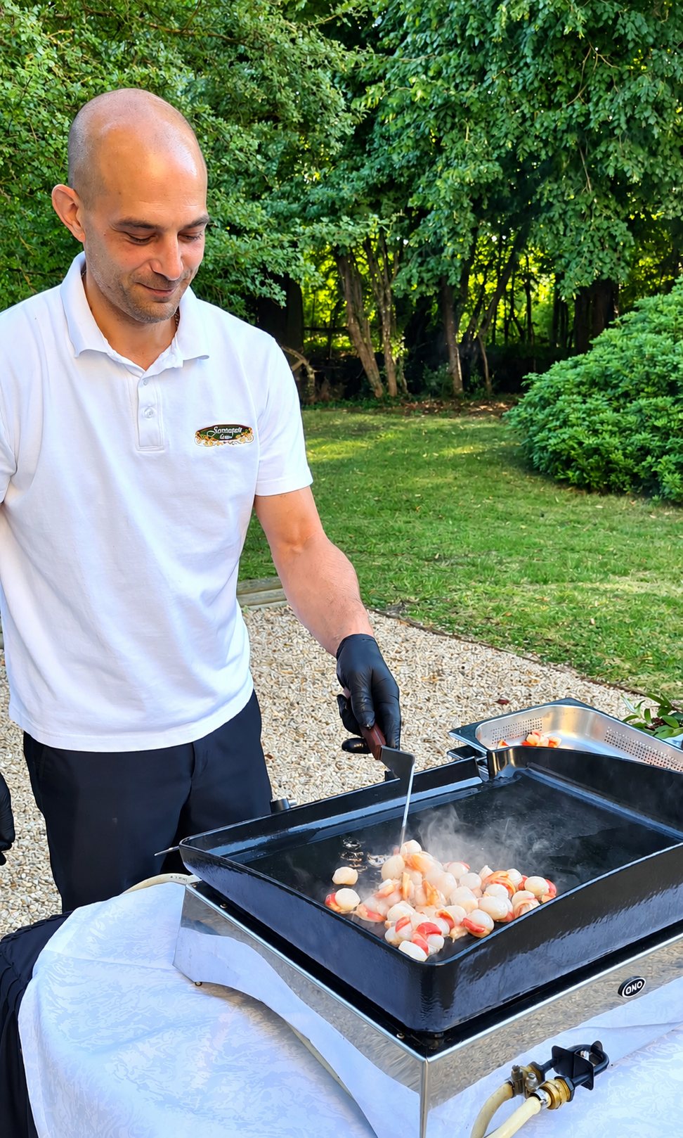 Un homme en polo blanc et gant noir fait cuire des noix de Saint-Jacques sur une plancha posée sur une table dans un jardin.
