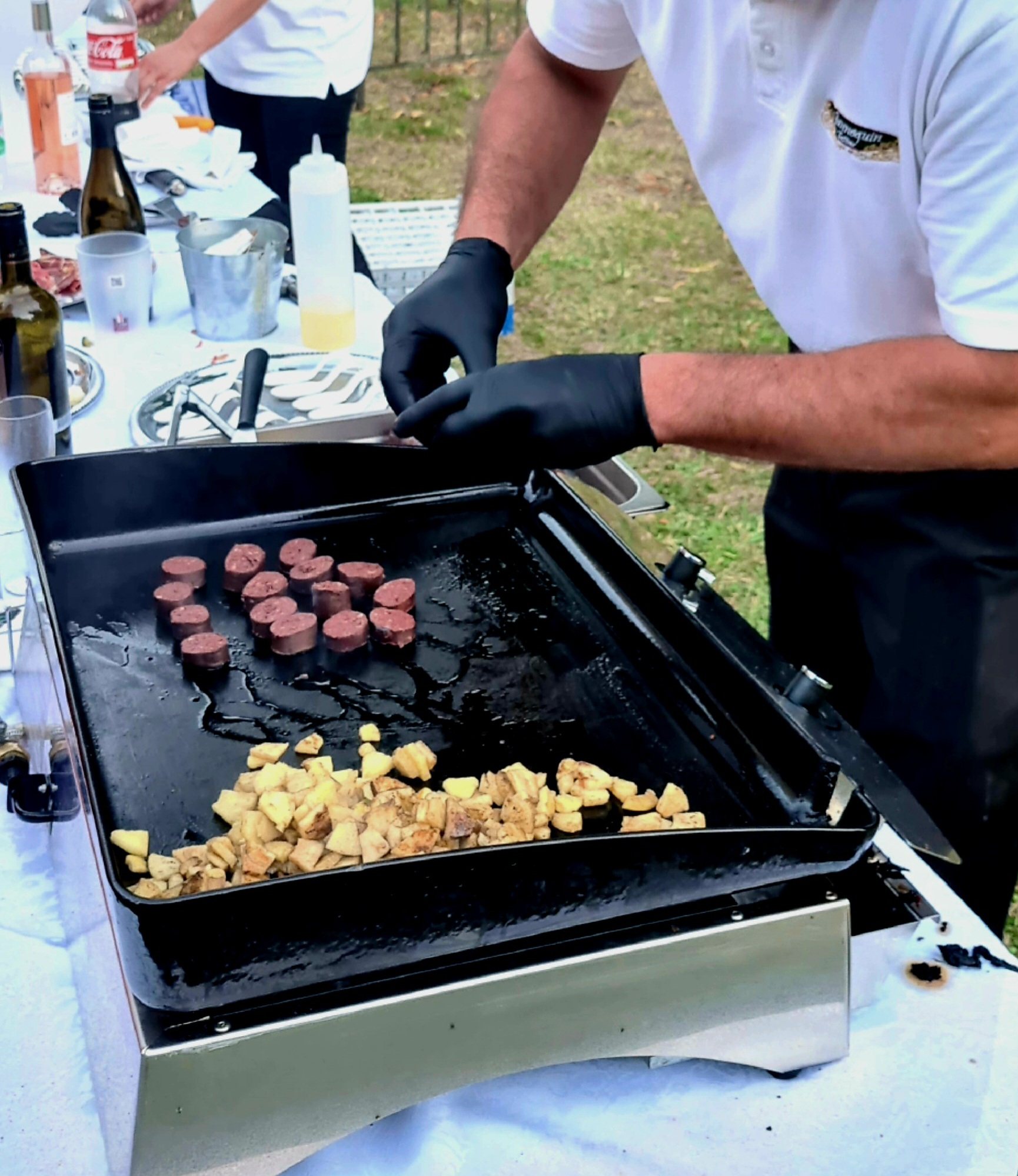 Un cuisinier en gants noirs fait cuire des rondelles de saucisse et des cubes de pommes de terre sur une plancha posée sur une table dehors.