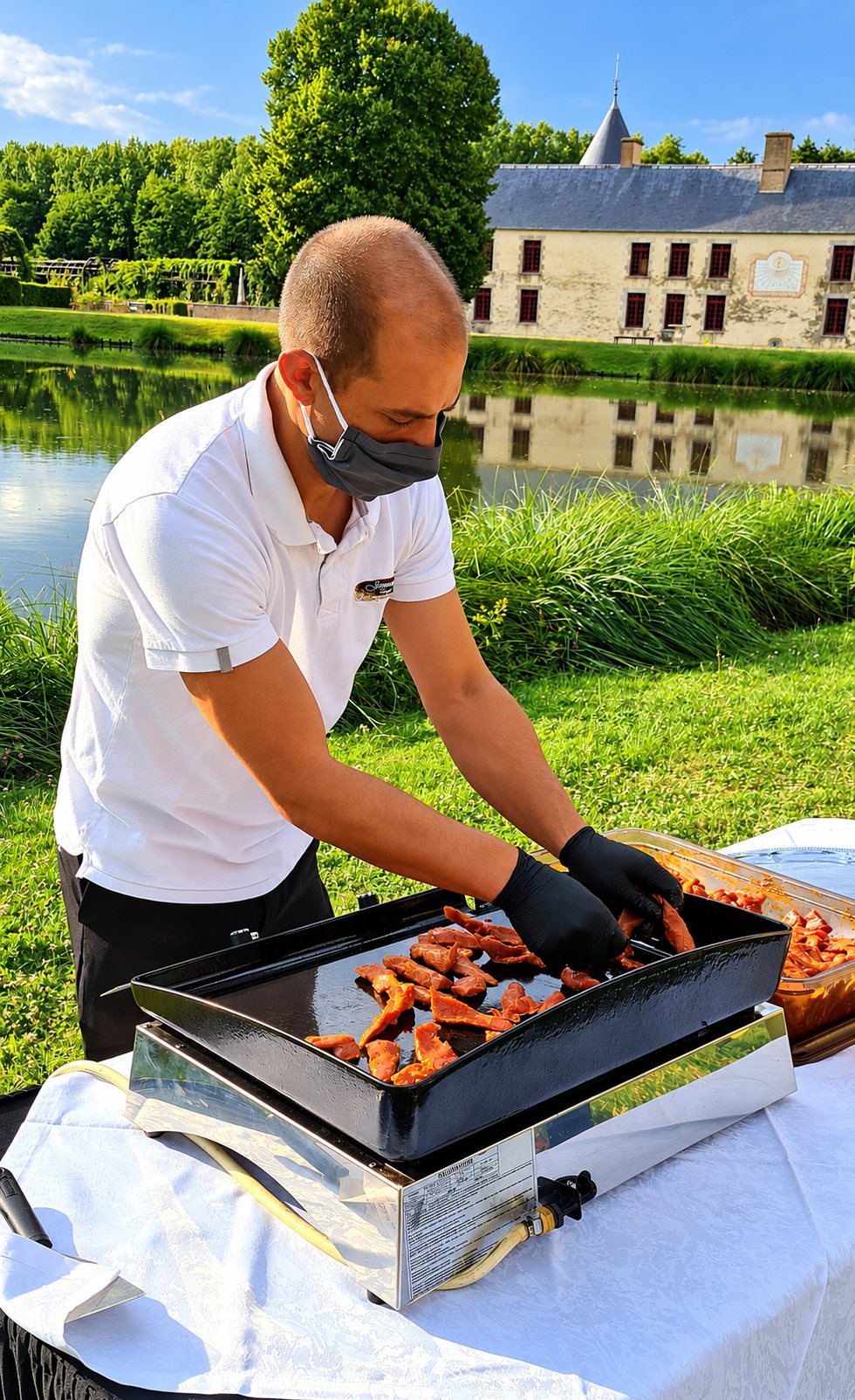 Un homme en polo blanc et masque fait cuire des morceaux de viande marinée sur une plancha posée sur une table, devant un étang et un château.