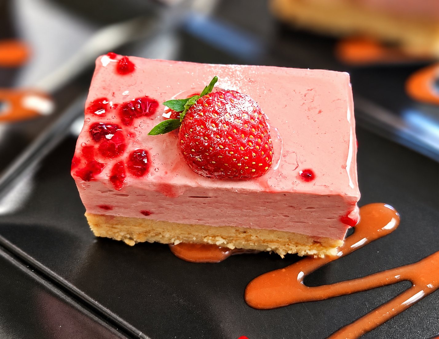 Une part rectangulaire d'entremets rose surmontée d'une fraise fraîche, posée sur une assiette noire avec un trait de coulis de fruits.