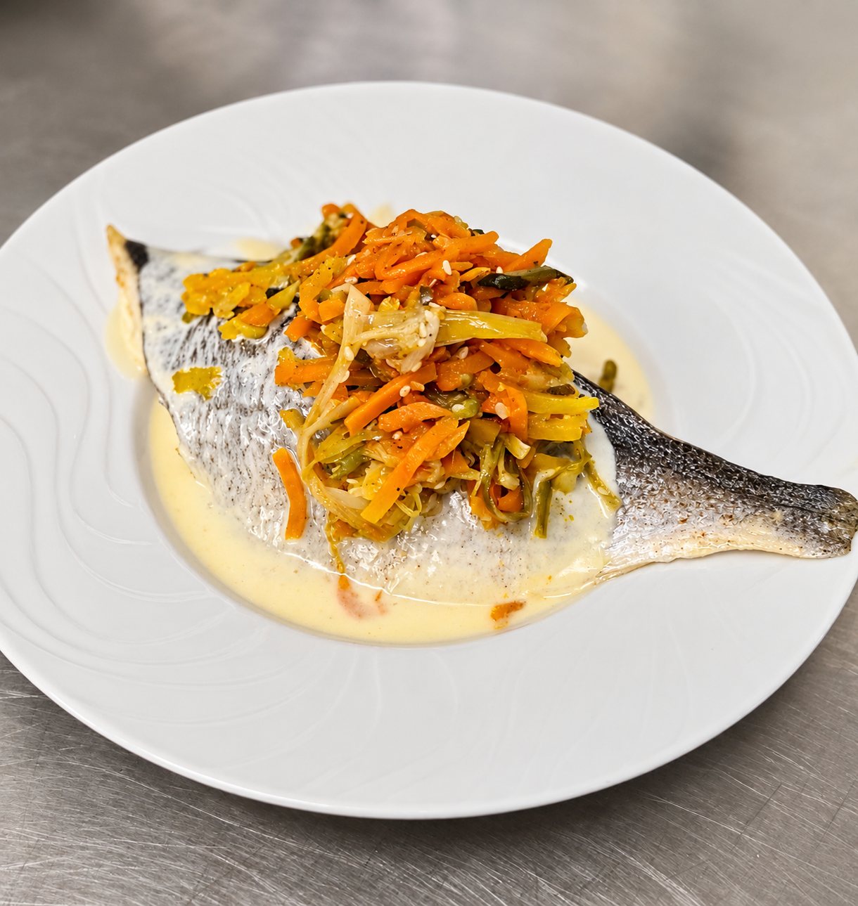 Un filet de poisson à la peau croustillante, nappé de sauce crémeuse et garni d'une julienne de carottes et de poireaux, servi dans une assiette creuse blanche posée sur un plan de travail en inox.