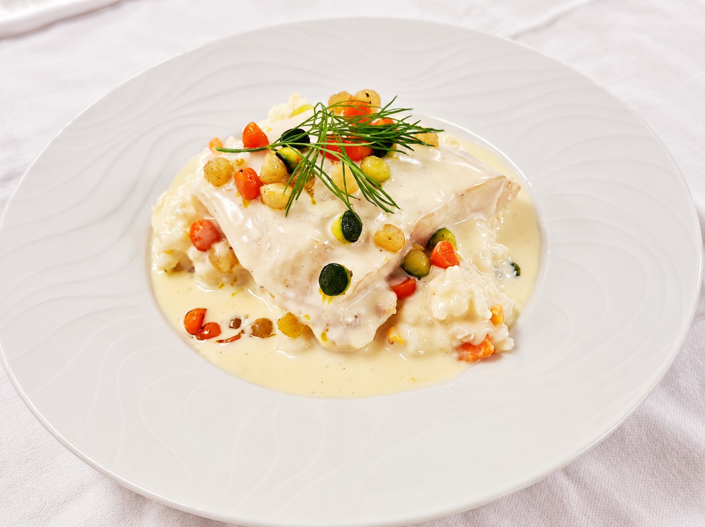 Une assiette creuse blanche contient un filet de poisson nappé de sauce crémeuse, posé sur du risotto avec des dés de carottes et de courgettes et une branche d'aneth.