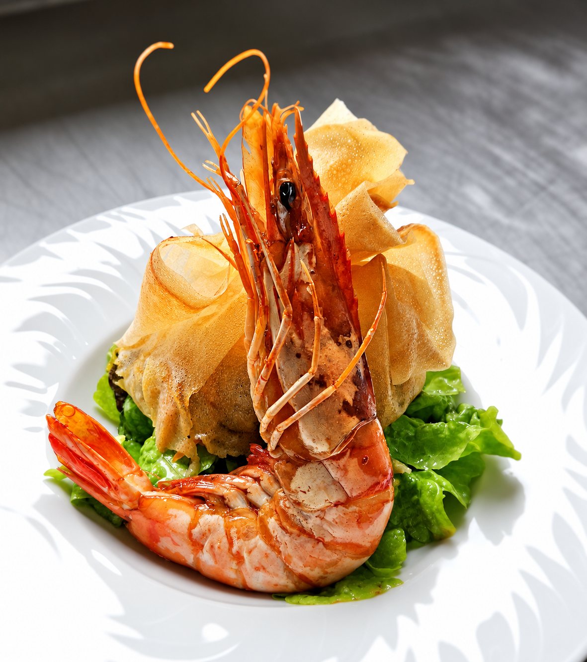 Une grosse gambas rôtie posée devant une aumônière de pâte croustillante sur un lit de salade verte, dans une assiette blanche.