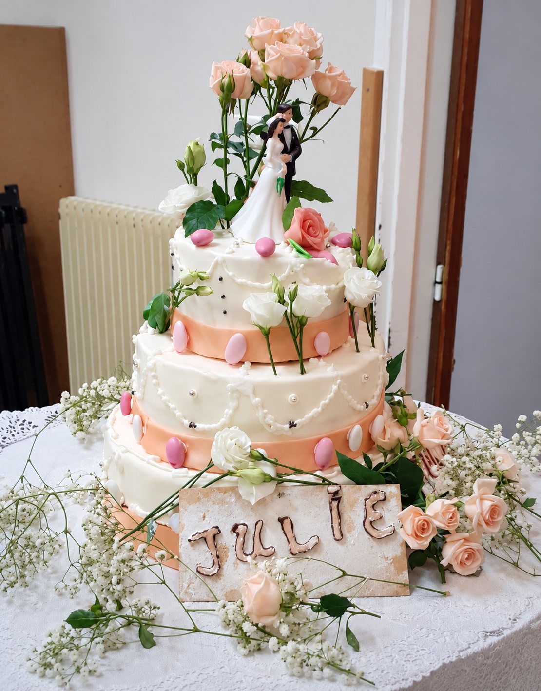 Gâteau de mariage blanc et pêche à trois étages, orné de roses, de gypsophile et de dragées roses, surmonté d'un couple de mariés, avec une pancarte portant le prénom Julie.