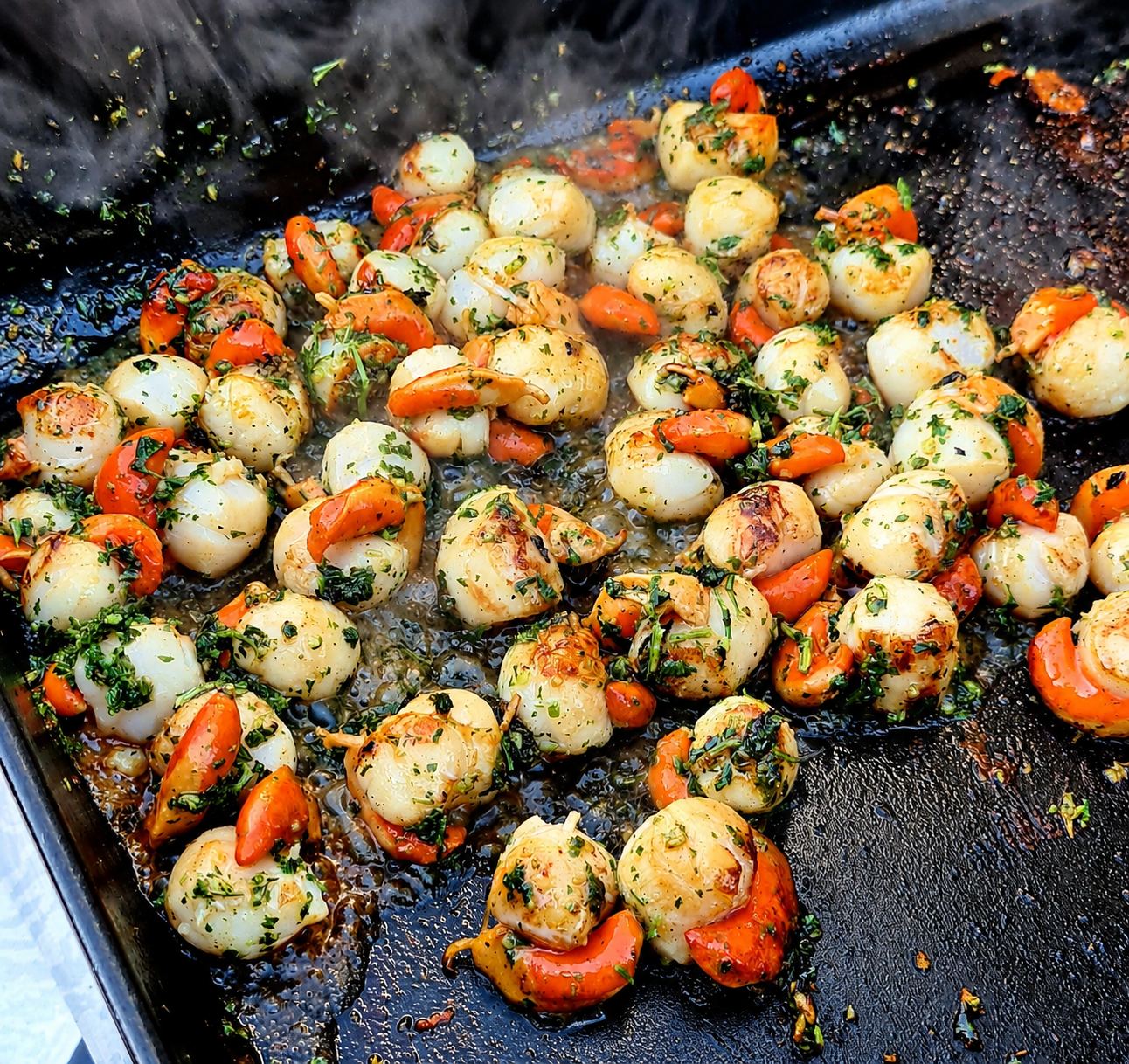Des noix de Saint-Jacques avec leur corail orangé, parsemées de persil et d'ail, cuisent sur une plancha noire d'où s'échappe de la vapeur.