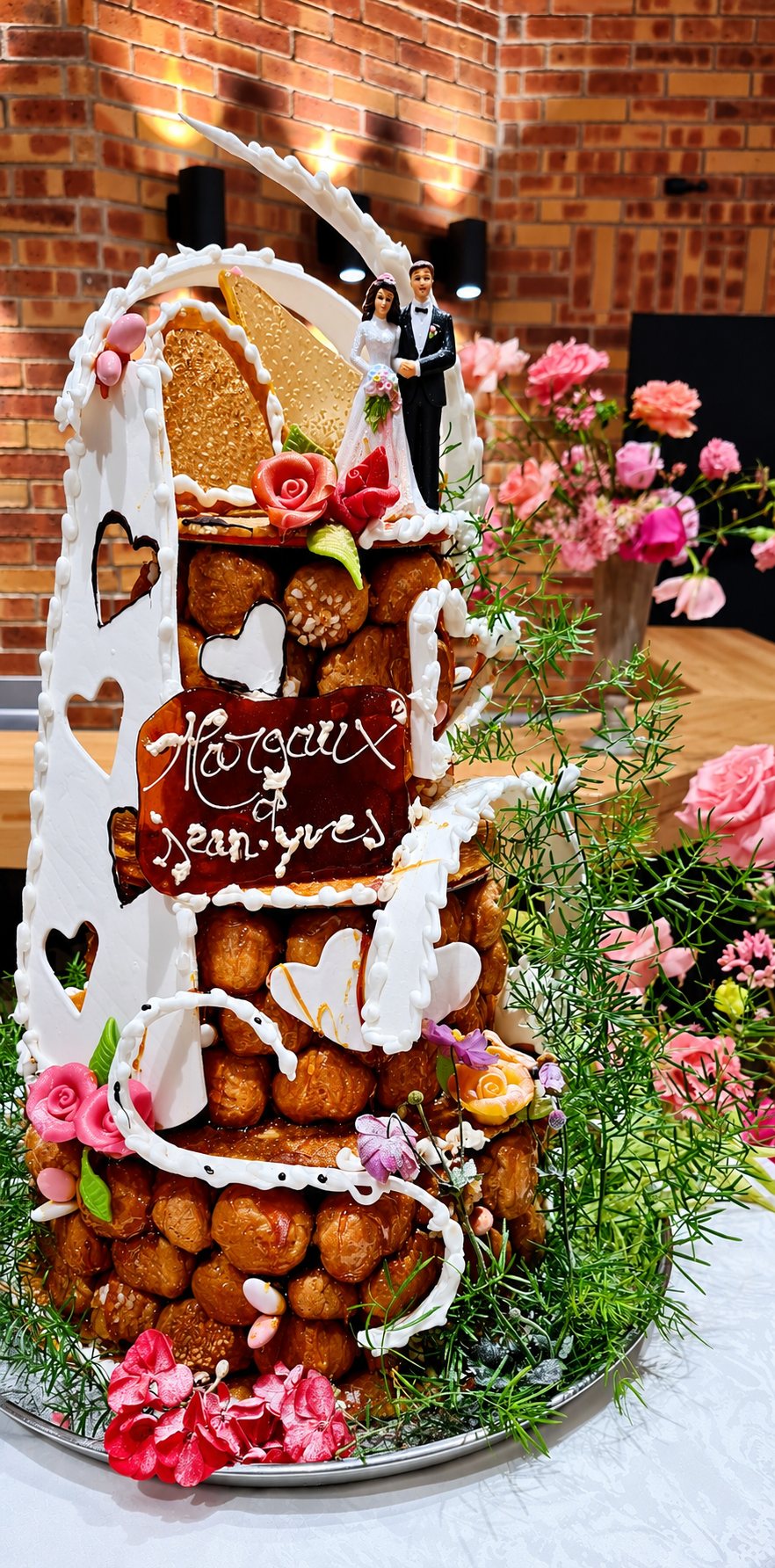 Pièce montée de mariage en choux, ornée de glaçage blanc, de fleurs roses et de figurines de mariés, posée sur une table devant un mur de briques.