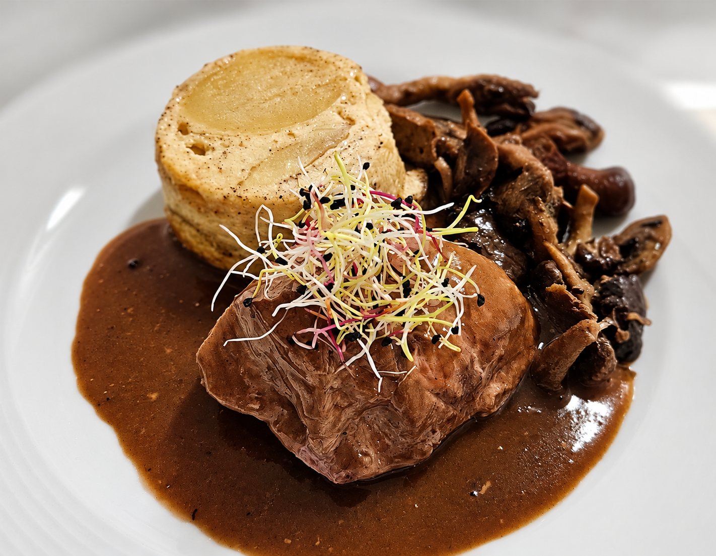 Assiette blanche garnie d'un morceau de viande nappé de sauce brune, accompagné de champignons et d'un petit flan, décoré de germes colorés.