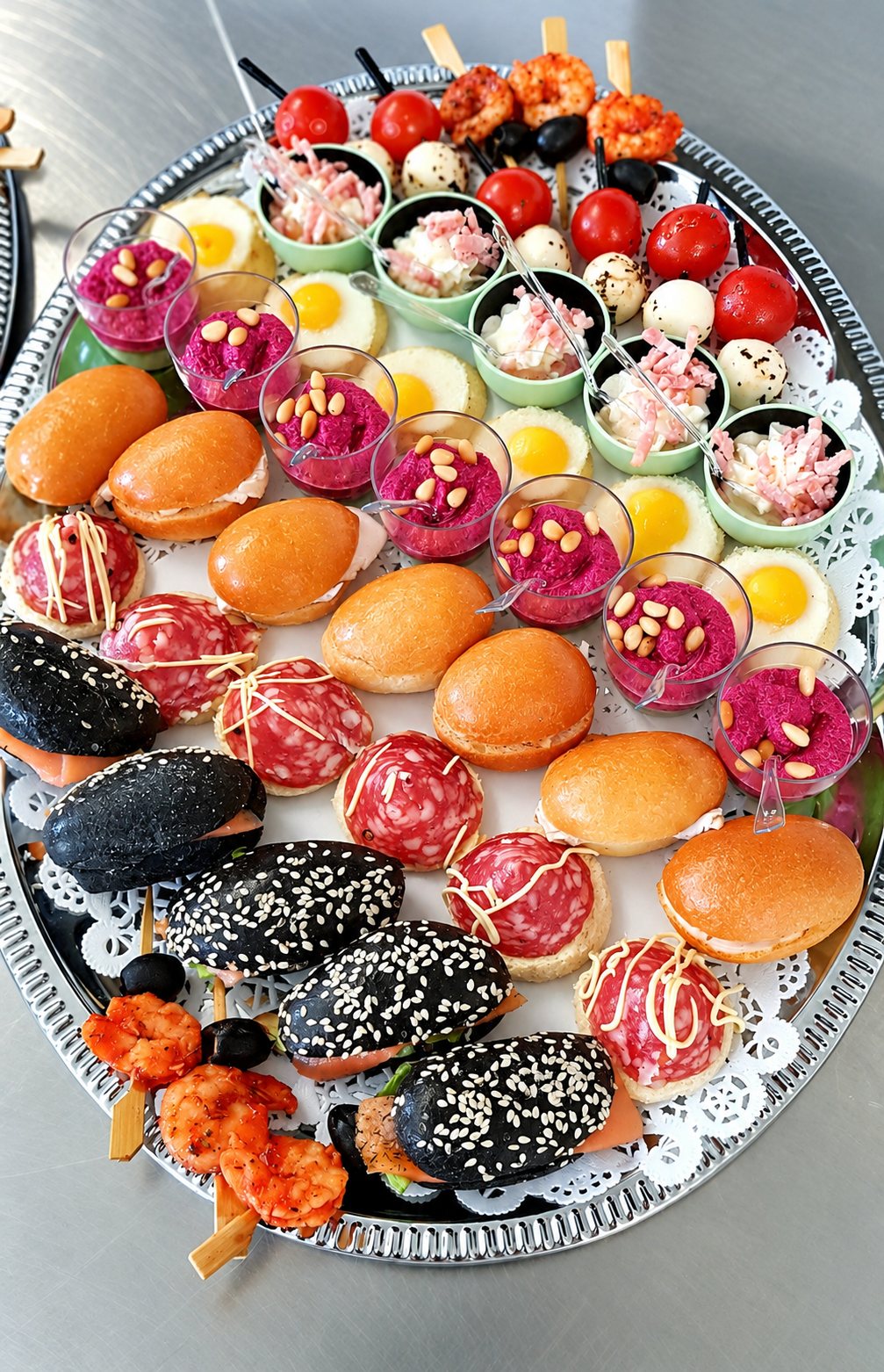 Un plateau argenté ovale garni de mini burgers noirs et dorés, de canapés au salami, de verrines de houmous de betterave aux pignons, d'œufs au plat miniatures et de brochettes de crevettes.