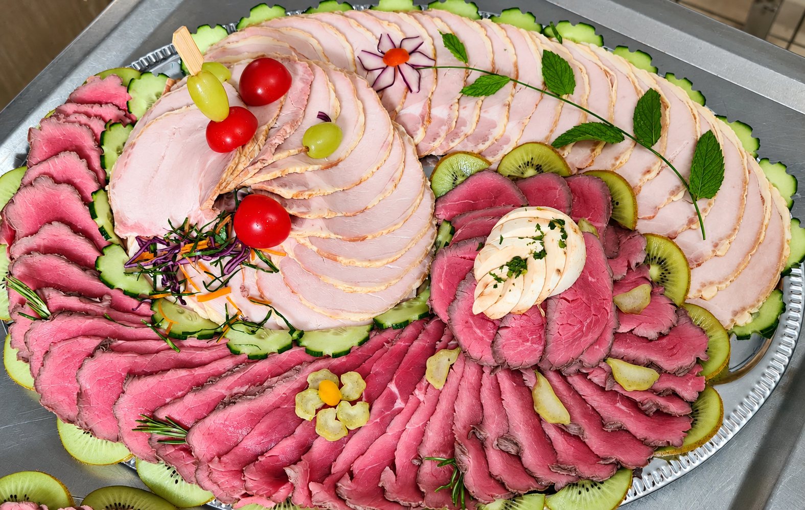 Un grand plateau de service garni de tranches de rôti de porc et de rôti de bœuf rosé, décoré de concombre, kiwi, tomates cerises, raisin et feuilles de menthe.
