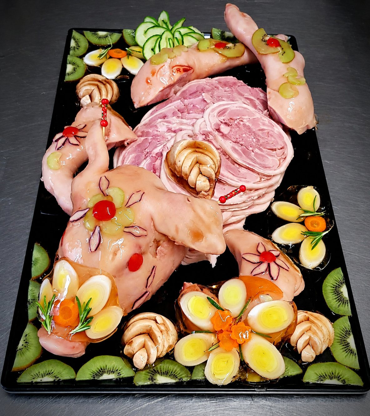 Un plateau noir présente une volaille décorée de fleurs en poireau et de cerises, entourée de tranches de jambon rosé, de kiwis et de concombre, le tout nappé de gelée.