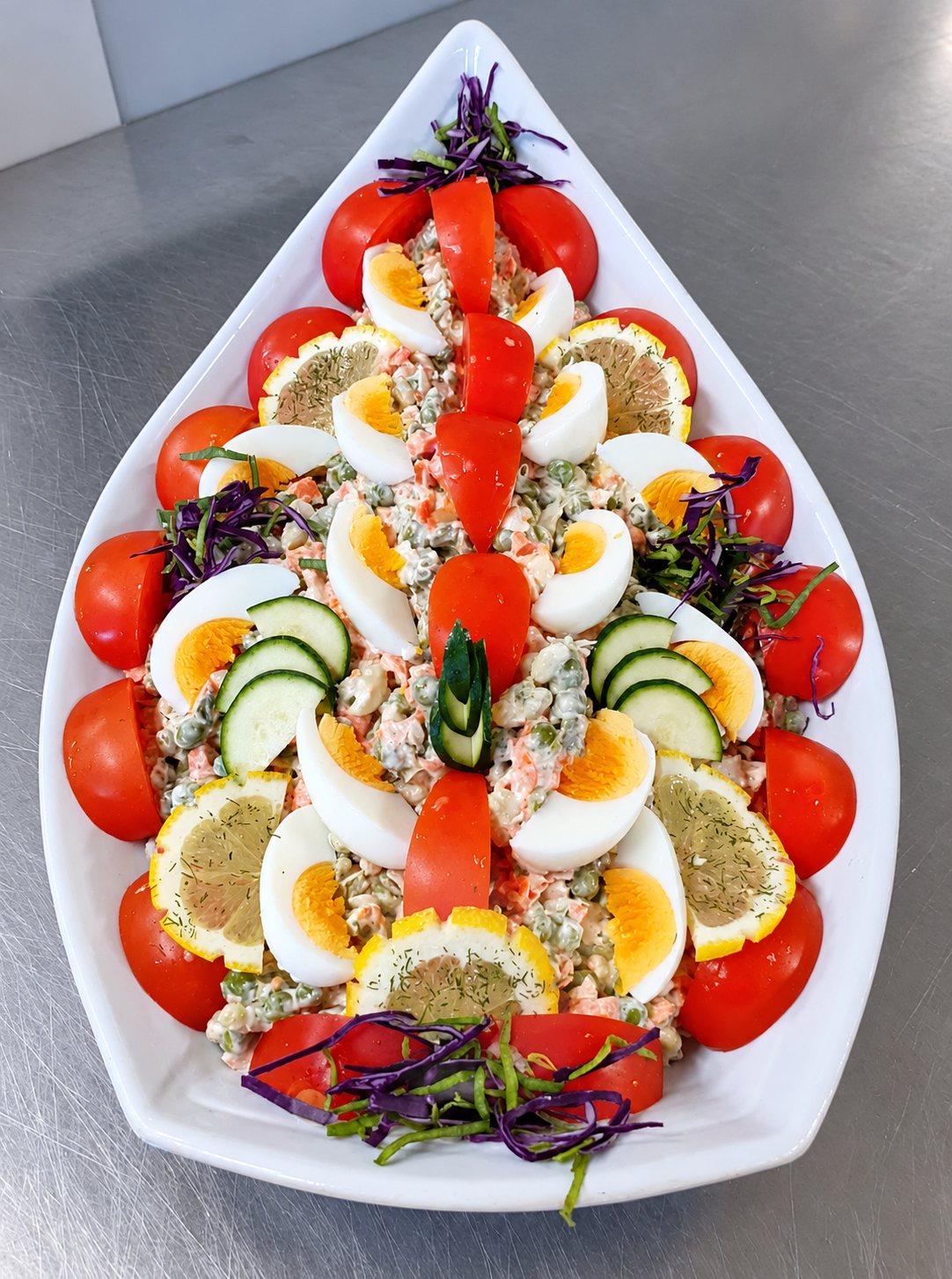 Plat blanc en forme de goutte garni d'une salade de légumes à la mayonnaise, décorée de demi-tomates, d'œufs durs, de rondelles de citron et de concombre.
