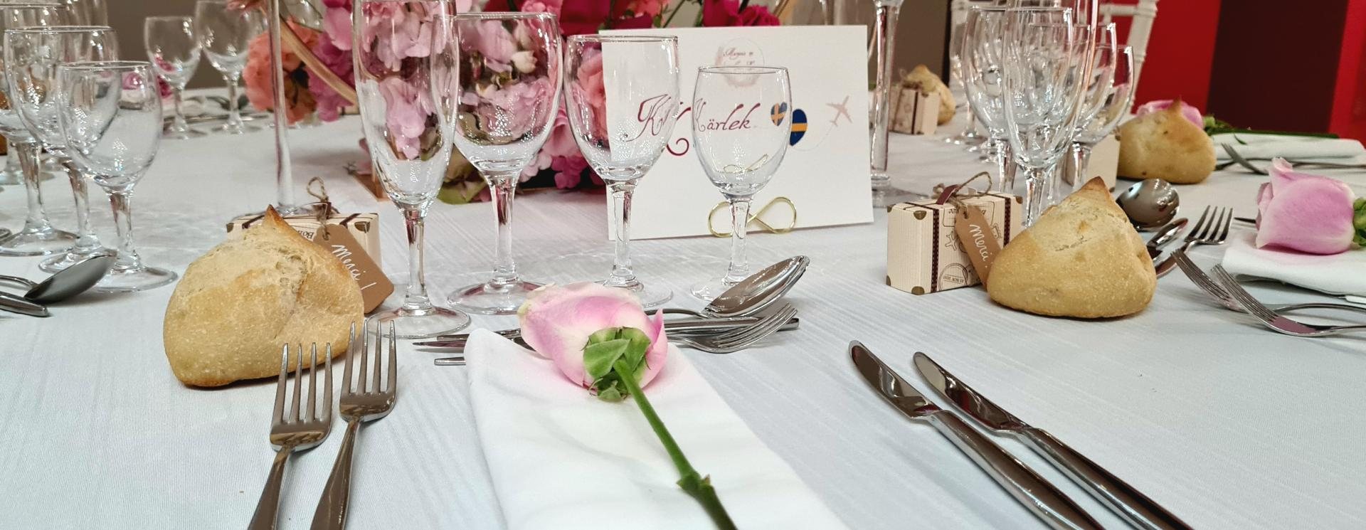 Table de réception à la nappe blanche dressée avec verres, couverts, rose rose sur une serviette, petits pains, boîtes à dragées et carton de place.