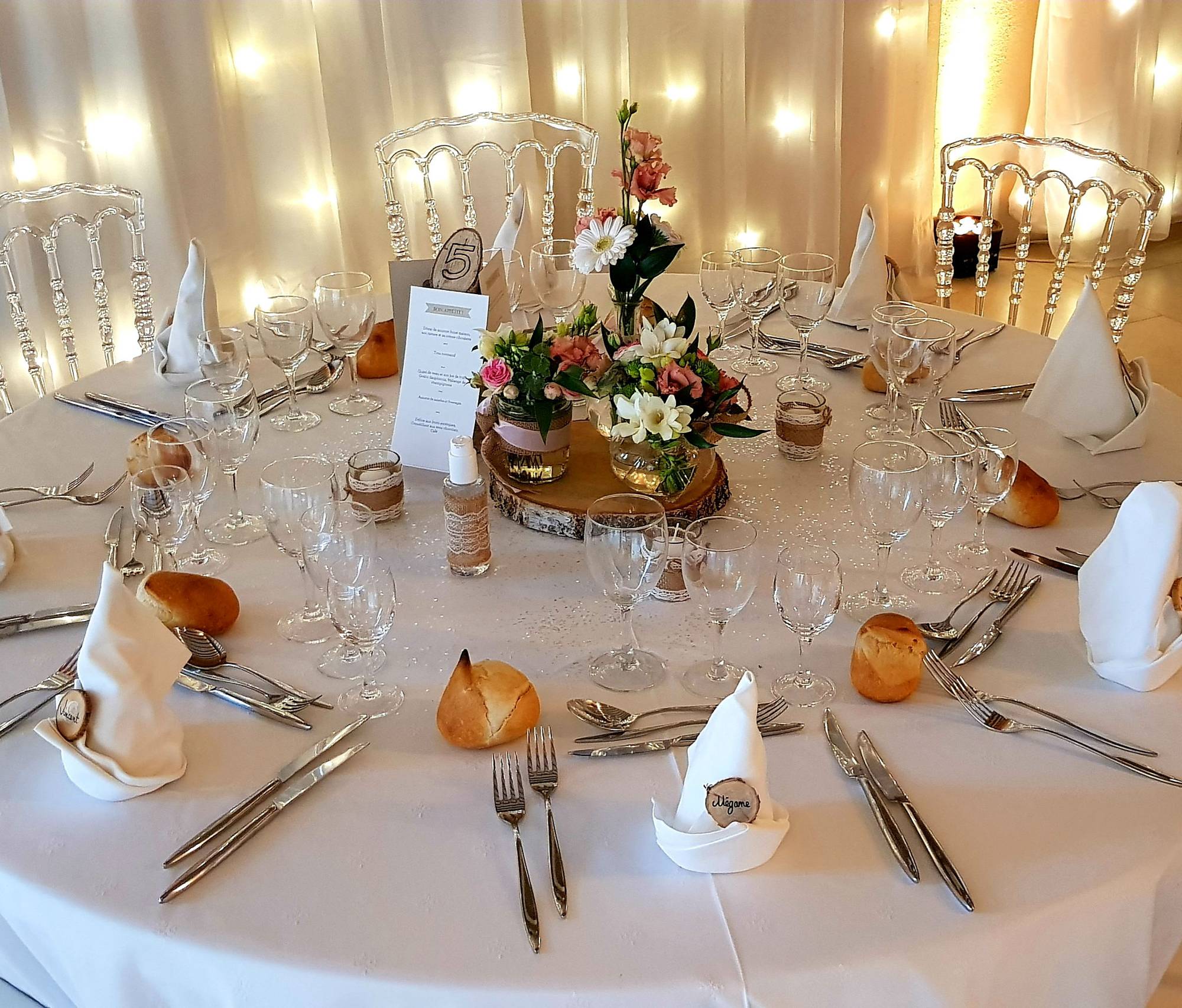 Table de réception ronde nappée de blanc avec verres, couverts, petits pains, menu et bouquets de fleurs sur une rondelle de bois, devant des rideaux illuminés.