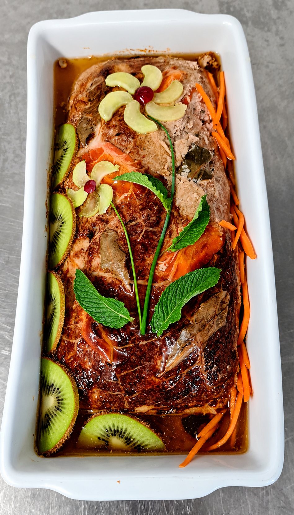 Terrine de viande dans un plat blanc rectangulaire, décorée de feuilles de menthe, de fleurs stylisées, de carottes râpées et de tranches de kiwi.