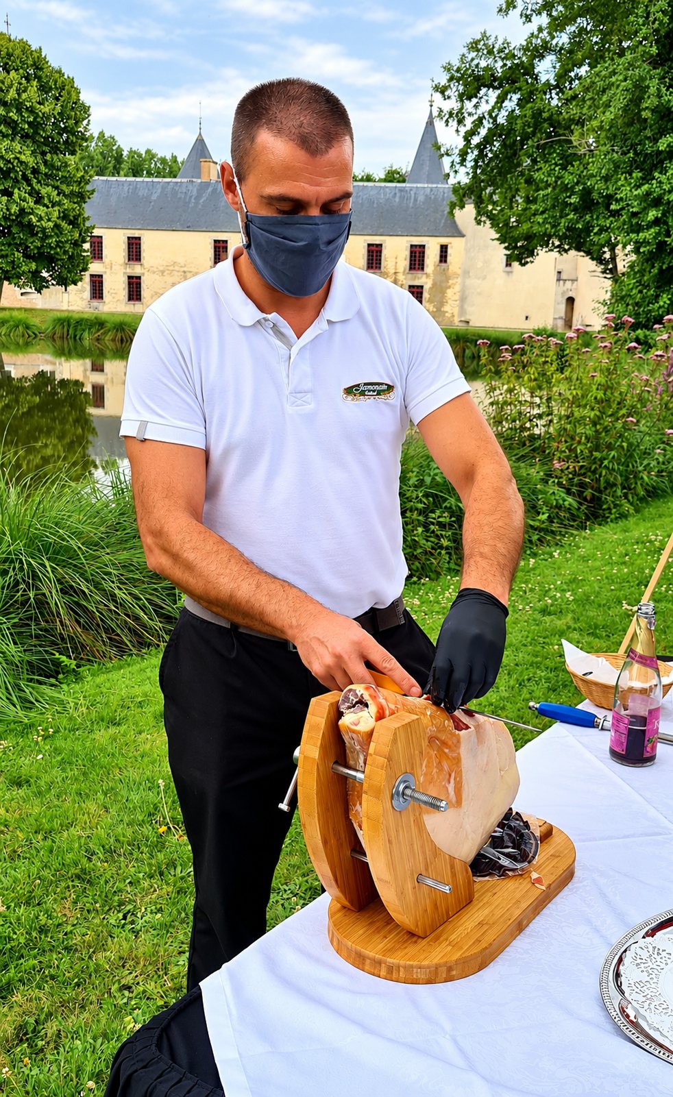Un homme masqué en polo blanc découpe un jambon fixé sur un support en bois, sur une table dressée dans un parc devant un château et un plan d'eau.