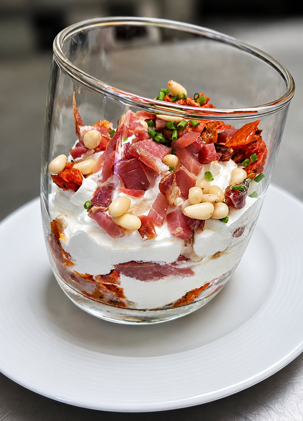 Verrine en verre posée sur une assiette blanche, garnie de crème, de jambon cru, de pignons de pin, de tomates séchées et de ciboulette.